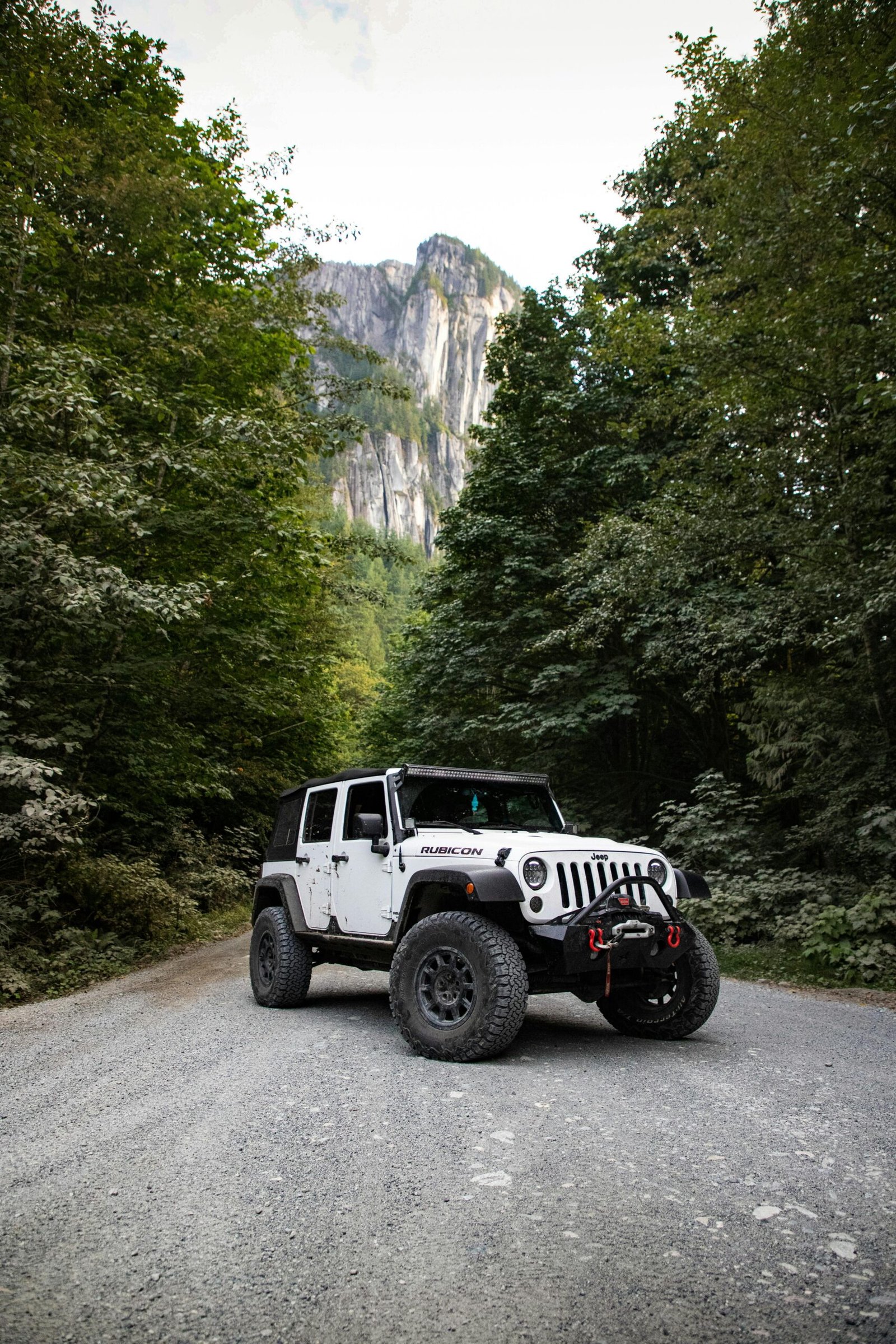 A rugged Jeep Wrangler on a gravel road surrounded by lush forest and towering mountains.