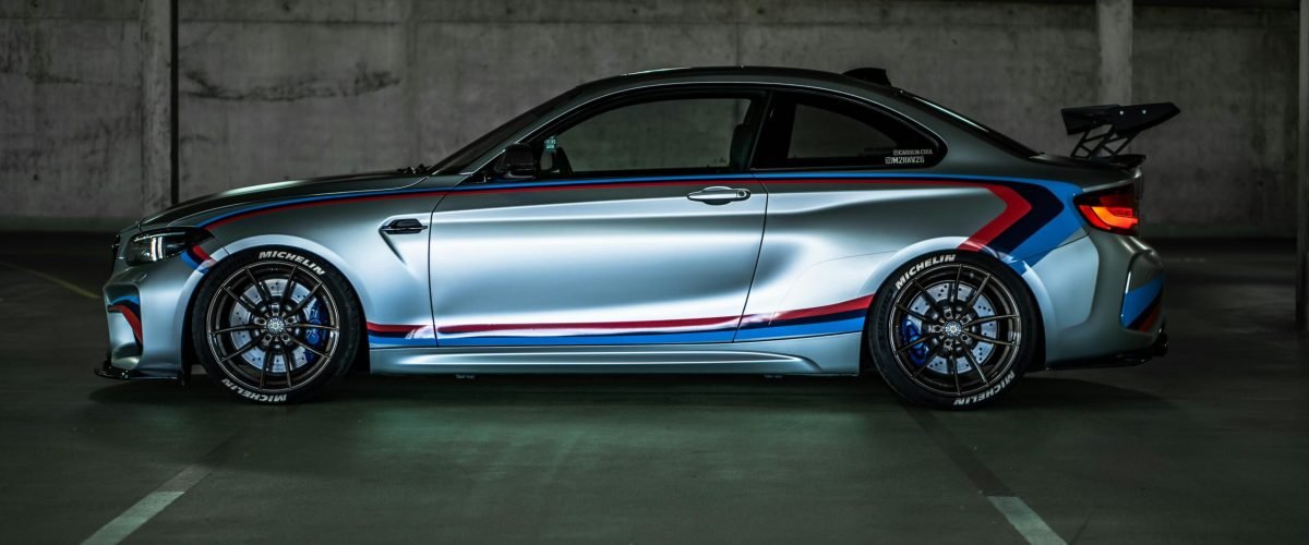 Side view of a BMW M2 sports car with racing stripes, parked in a dimly lit garage.